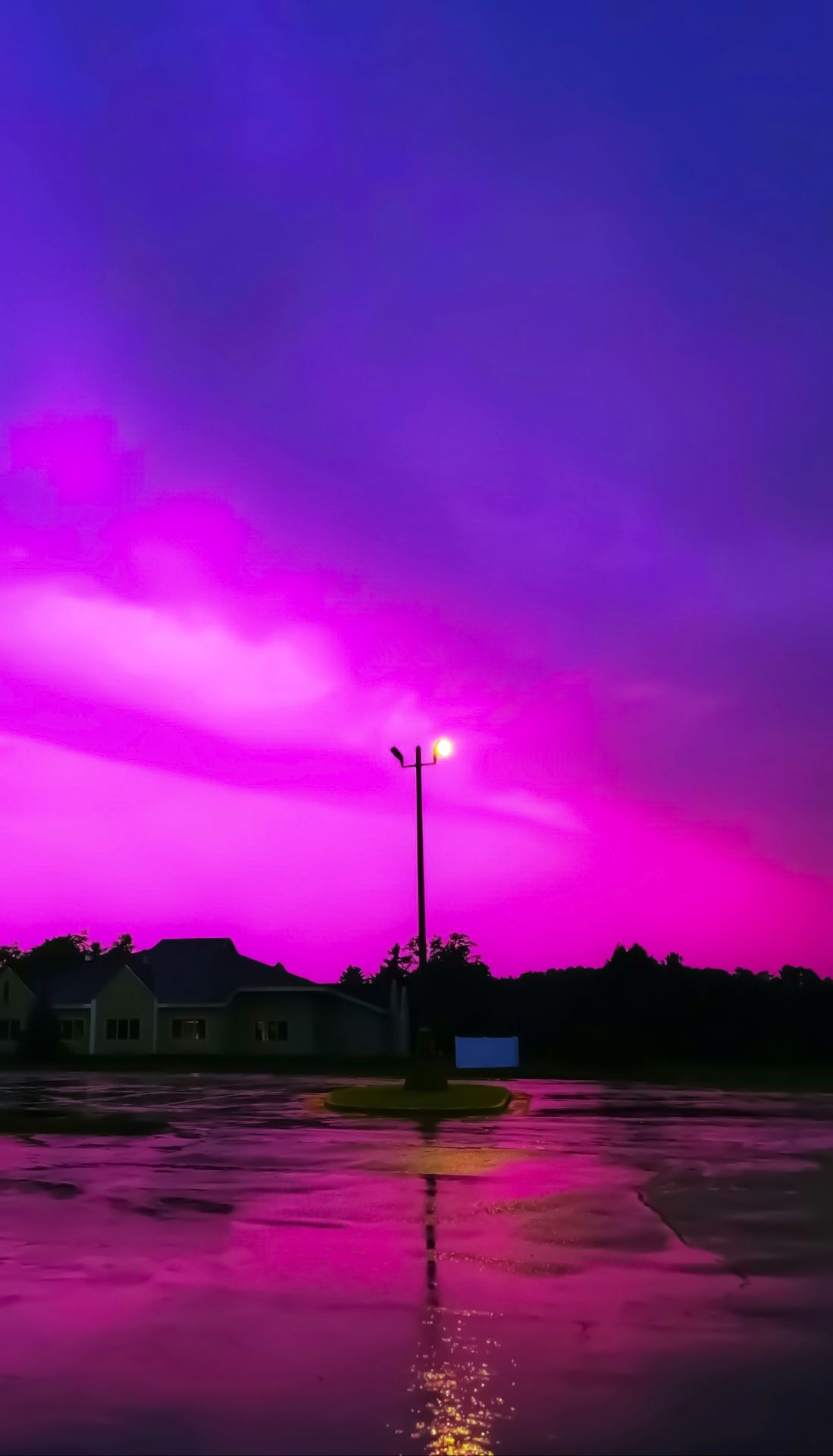 A glowing streetlamp illuminates a wet parking lot under a surreal, vibrant magenta and purple sky, creating a dreamlike, liminal atmosphere.