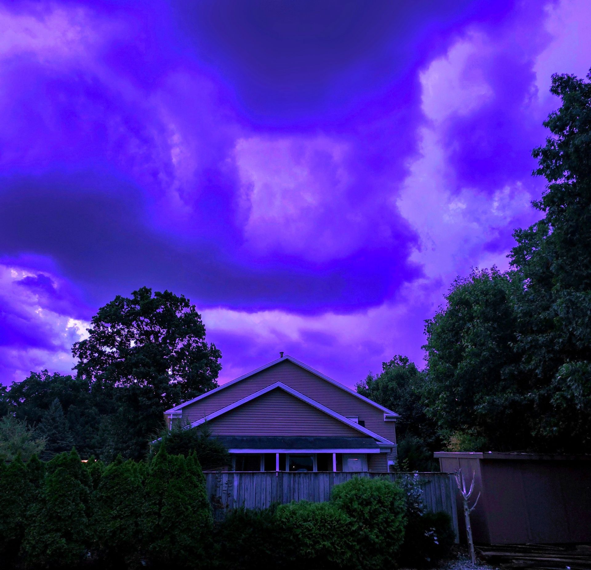 A suburban house sits beneath a surreal, vibrant purple storm sky, creating an ethereal, liminal atmosphere framed by dark, silhouetted trees and foliage.