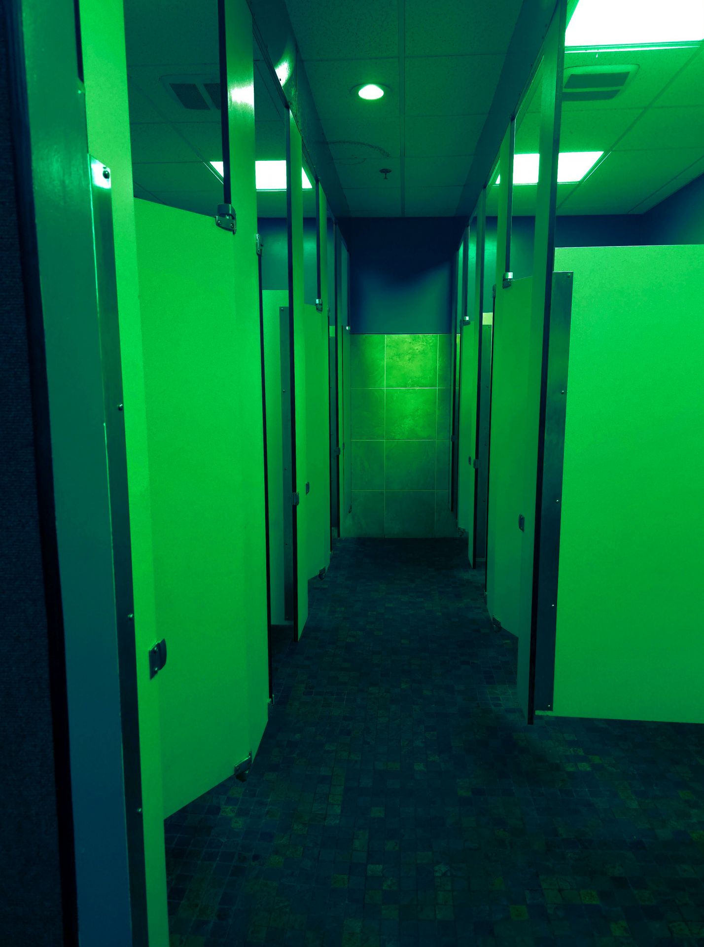 A symmetrical, liminal view down a public restroom aisle bathed in eerie, vibrant green light, featuring rows of stall doors and tiled flooring.