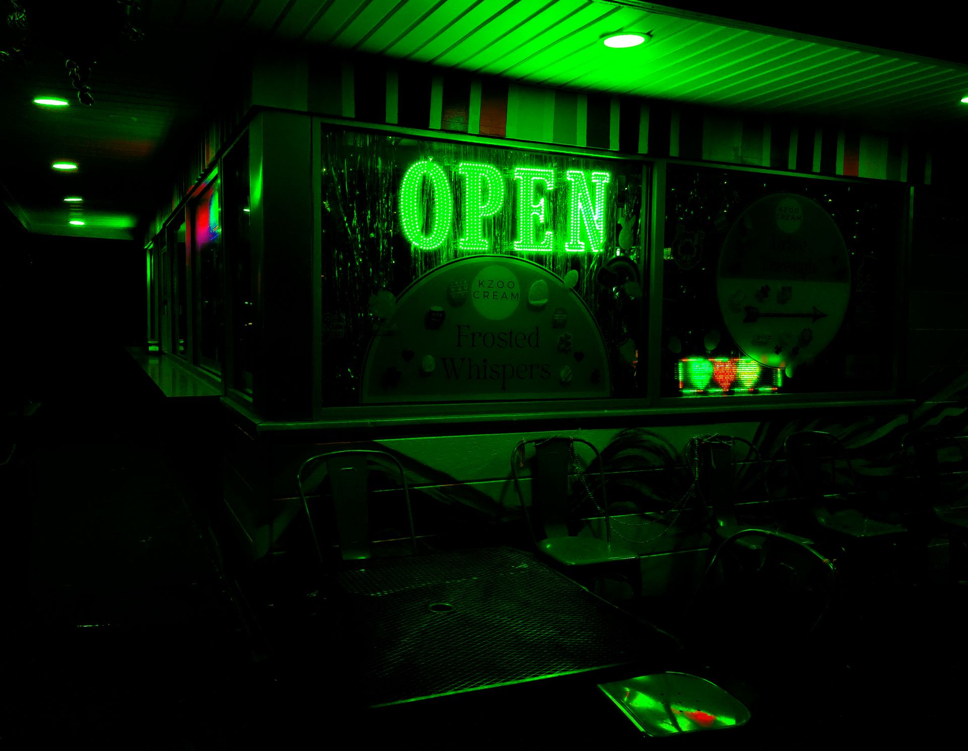 An ethereal, liminal night shot of a Kzoo Cream shop exterior, bathed in intense green neon light, featuring empty metal tables and chairs.