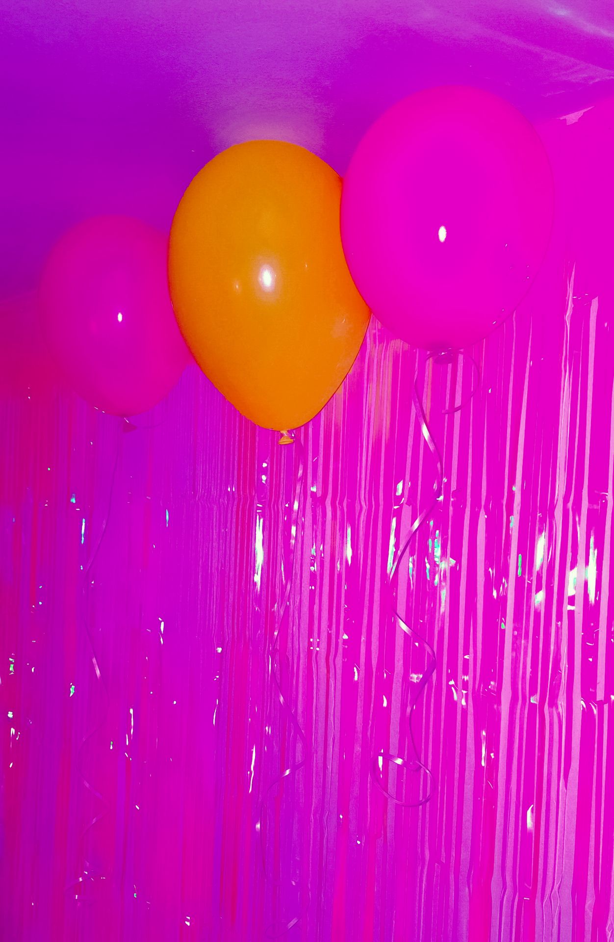 Three balloons—two pink, one orange—float against a shimmering, monochromatic magenta tinsel backdrop, creating a surreal, dream-like, and liminal party atmosphere.