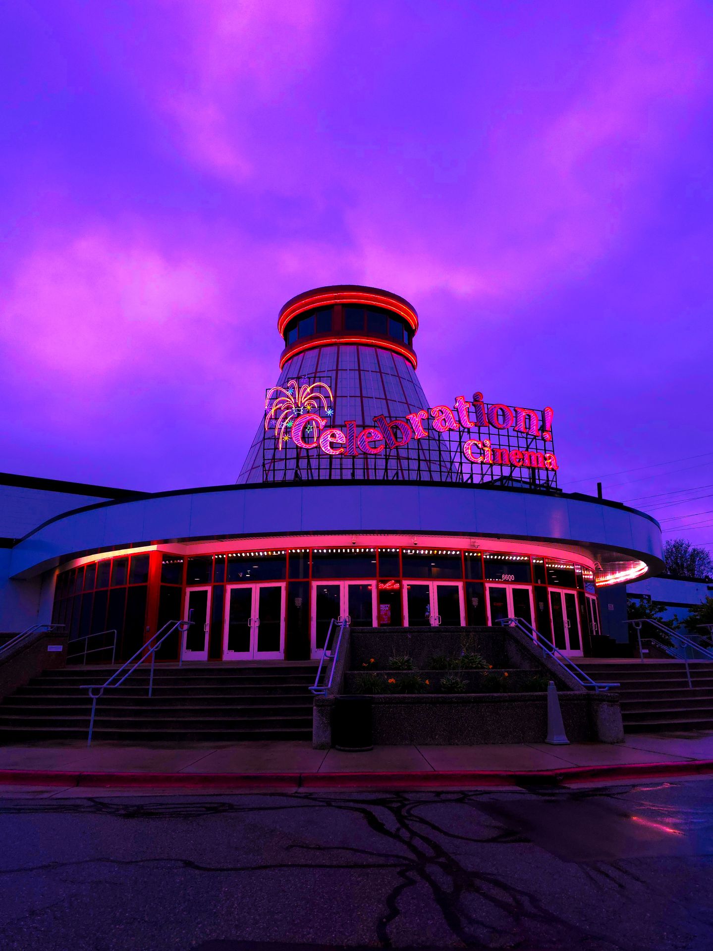 A neon-lit Celebration! Cinema entrance stands under a dreamlike, vibrant purple sky, featuring a conical tower and symmetrical stairs in a liminal atmosphere.