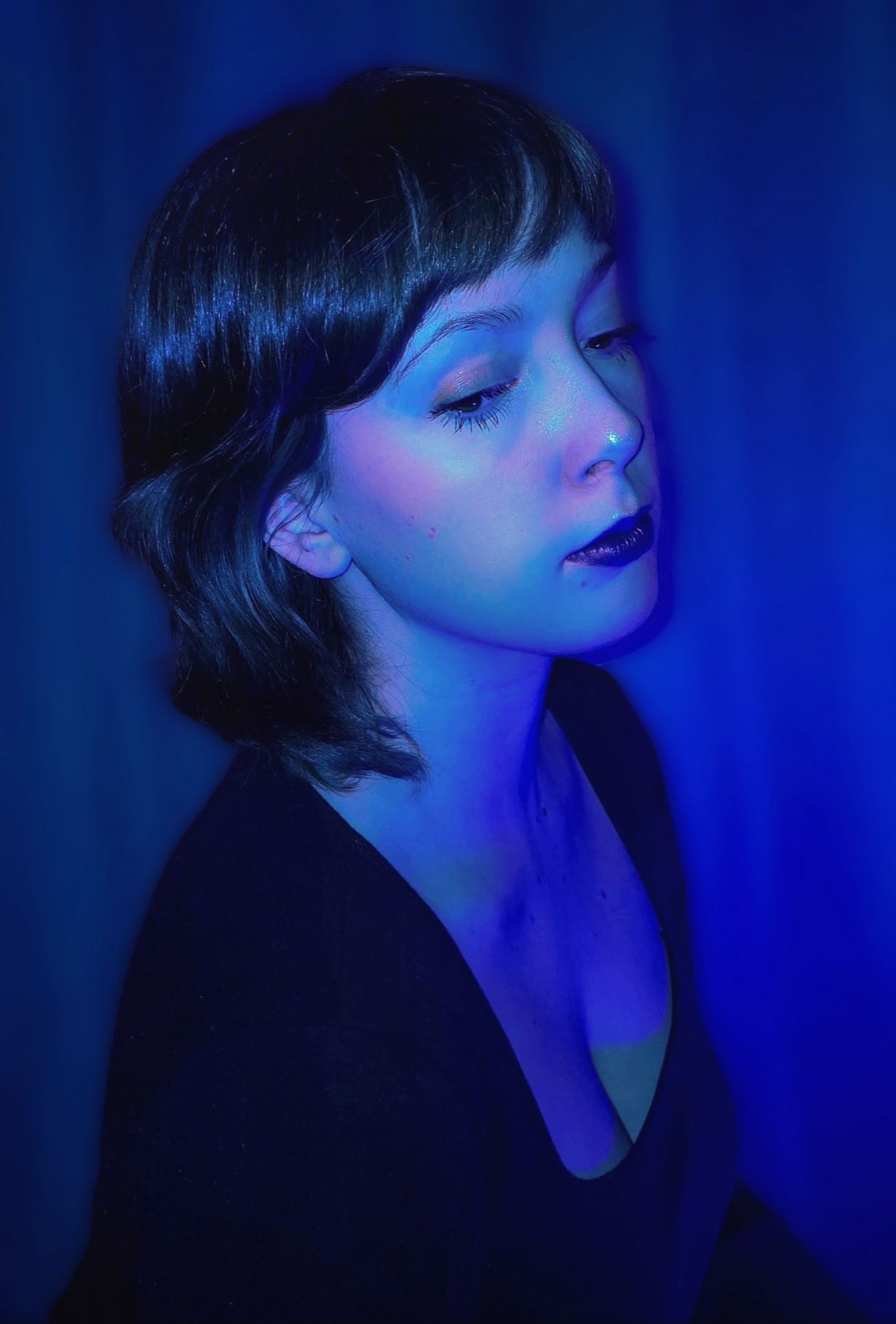 A woman with dark hair and dark lipstick gazes downward in a dream-like, ethereal portrait bathed in moody, saturated blue studio lighting.