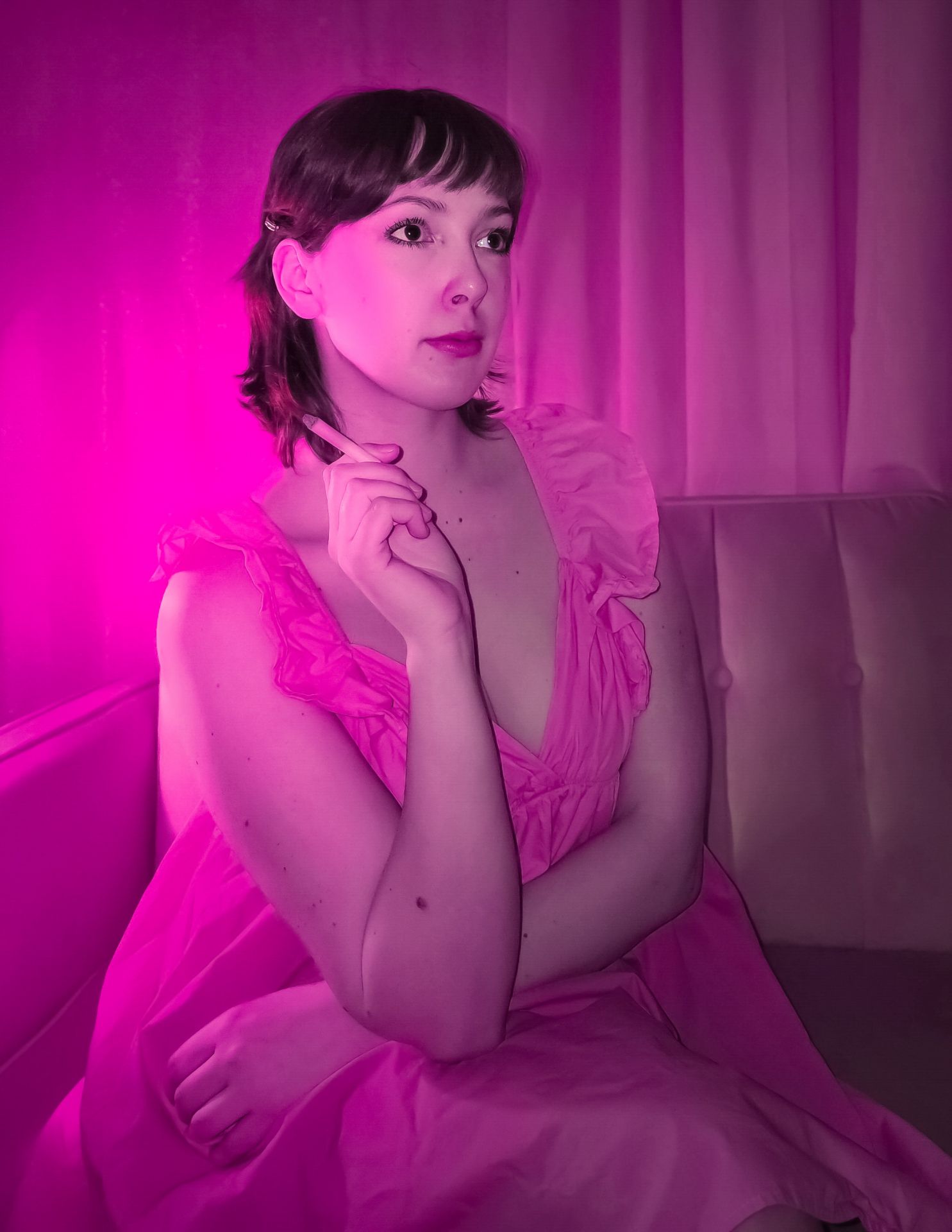 A woman in a ruffled pink dress sits on a couch, bathed in ethereal, dream-like neon pink light, creating a surreal, liminal atmosphere.