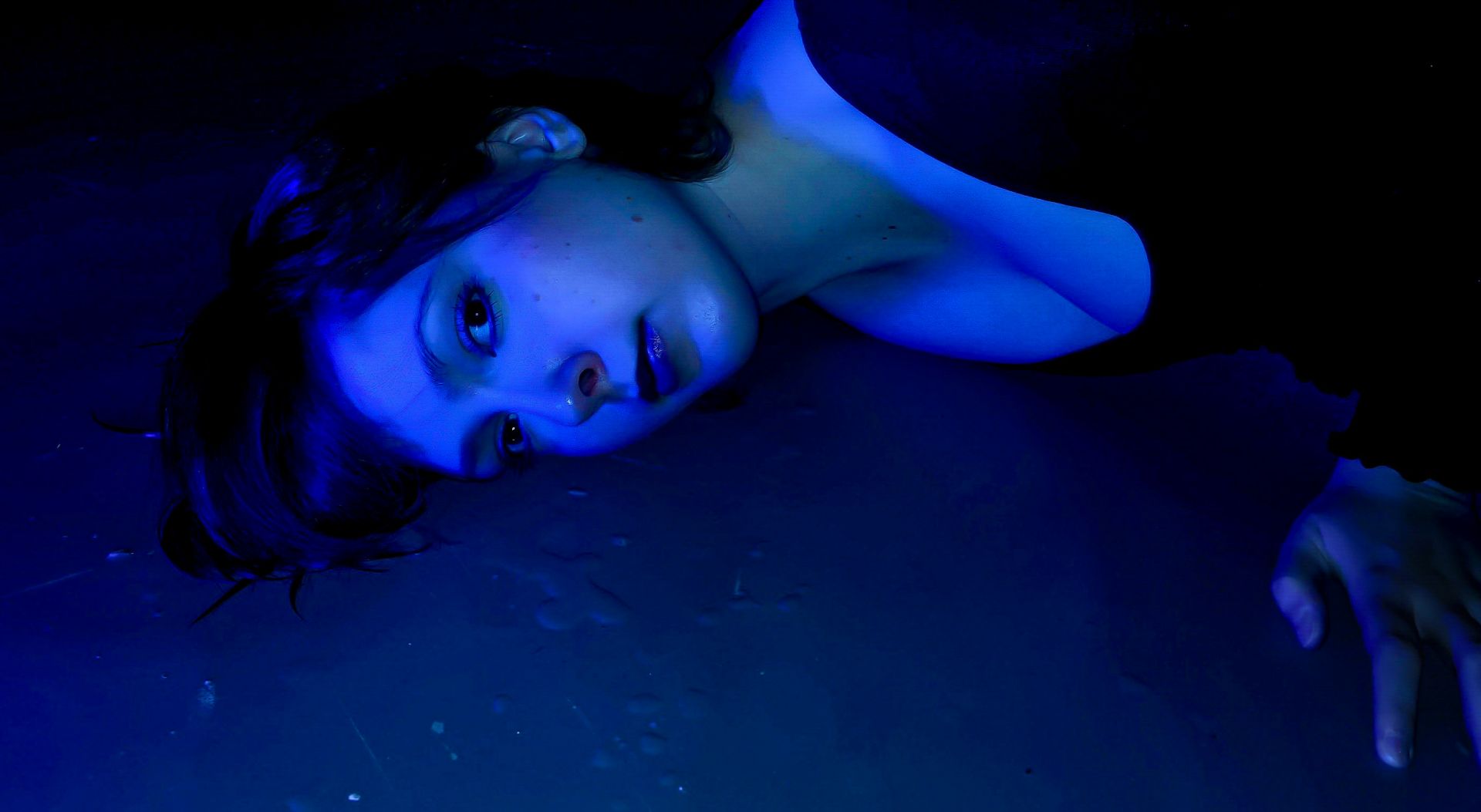 A woman in a black top lies on a dark surface, illuminated by ethereal blue light, creating a moody, dream-like, and liminal atmosphere.
